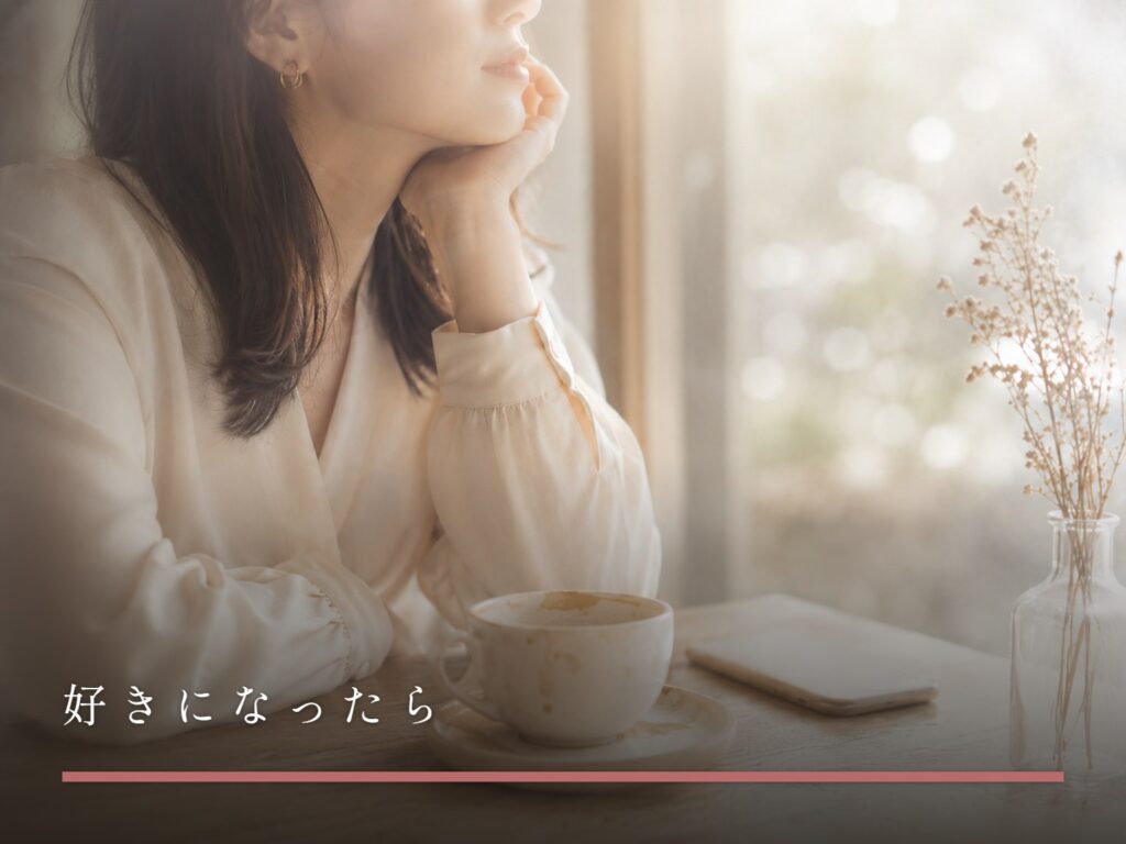 カフェの窓際で頬杖をつき外を眺める女性。テーブルにはコーヒーと画面を伏せたスマートフォン（好きになったら）