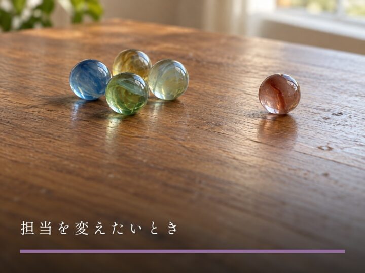 木の机の上で、ひとつだけ少し離れたところに転がった色違いのビー玉(担当を変えたいとき)